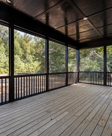 The image shows a spacious screened-in porch or lanai with a clean, modern look. It features light-colored wooden deck flooring, dark-painted railings, and full-height screen panels that wrap around the space, allowing an unobstructed view of the surrounding trees and greenery. The ceiling is finished in a dark tone with recessed lighting, creating contrast with the lighter floor. Natural light fills the area, giving it an open, airy feel that’s ideal for relaxing outdoors while being protected from insects and the elements.